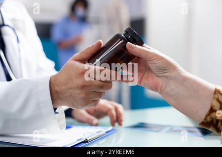 Großaufnahme von medizinischem Fachpersonal im Labormantel, der dem Patienten bei der Gesundheitscheckung die Pillen-Flasche präsentiert. Der Schwerpunkt liegt auf dem Behälter mit verschreibungspflichtigen Medikamenten mit Klemmbrett und Röntgenbild, die auf dem Tisch im Krankenhauszimmer sichtbar sind. Stockfoto