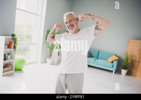 Energiegeladener Seniorenmann, der seine Muskeln mit Selbstvertrauen in einem hellen Innenraum mit aktivem Lebensstil und Fitness anspannt Stockfoto