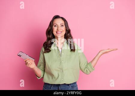 Foto einer fröhlichen schönen älteren Verkäuferin im Hemd hält Hand Mobile App für Miethäuser isoliert auf rosafarbenem Hintergrund Stockfoto