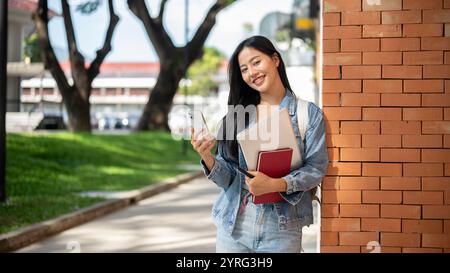 Eine attraktive asiatische Studentin in lässiger Kleidung, mit Rucksack und Büchern, steht an einer Ziegelwand und hält h Stockfoto