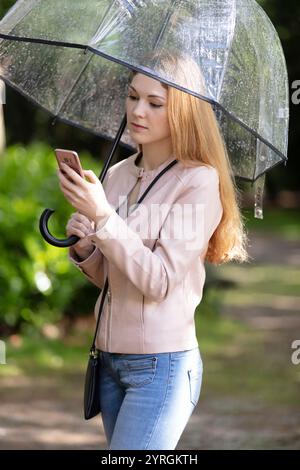 Schöne Frau mit transparentem Regenschirm, die unterwegs ist und sms liest Stockfoto