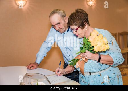 Ein älteres Ehepaar unterschreibt Dokumente in einem Standesamt in Russland, um ihren 50. Hochzeitstag zu feiern. Die Frau hält einen Blumenstrauß aus gelber Rose Stockfoto