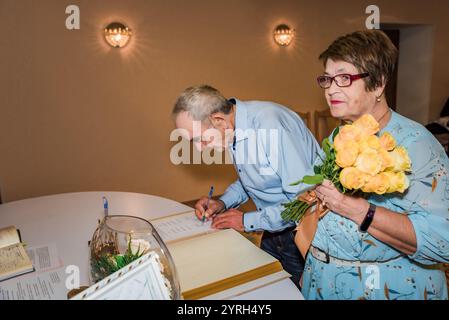 Ein älteres Ehepaar unterschreibt Dokumente in einem Standesamt in Russland, um ihren 50. Hochzeitstag zu feiern. Die Frau hält einen Blumenstrauß aus gelber Rose Stockfoto