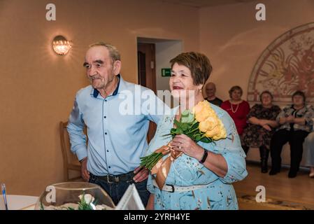 Ein Paar feiert seinen 50. Hochzeitstag (goldene Hochzeit) im Standesamt. Die Frau lächelt, während sie einen Blumenstrauß hält, und ihre Freunde Stockfoto