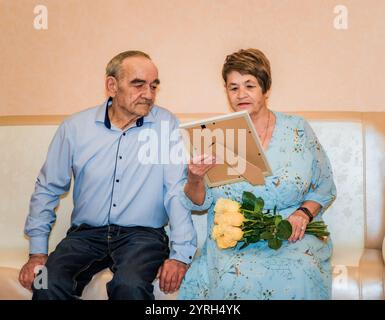 76-jähriger Mann und 71-jährige Frau feiern ihren 50. Hochzeitstag. Die Frau in einem blauen Kleid hält gelbe Rosen und sie sehen sich ein Geschenk an Stockfoto