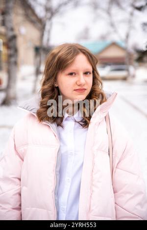 Die 15-jährige Russin in Winterjacke schaut direkt in die Kamera und steht vor schneebedeckten Birken mit Stadtkulisse. Ihr natürliches Stockfoto