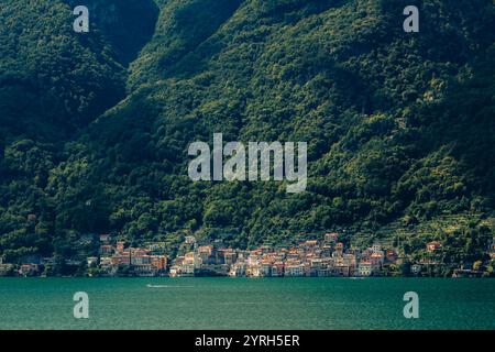 Das ruhige türkisfarbene Wasser des comer Sees spiegelt ein bezauberndes Dorf am Fuße einer grünen Bergkette wider und schafft ein fesselndes Panorama Stockfoto