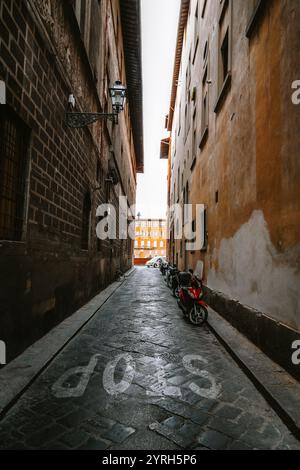 Geparkte Motorroller säumen eine enge Kopfsteinpflasterallee in florenz, italien, die zu einer größeren, helleren Straße und alten Gebäuden führt Stockfoto