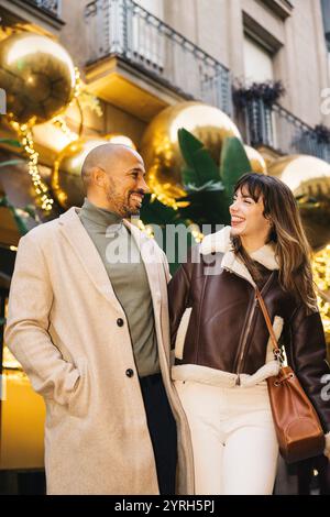 Ein lächelndes Paar genießt einen festlichen Einkaufstag in der Stadt, umgeben von funkelnden Dekorationen und goldenen Ornamenten. Ihr Lachen und ihre Freude erlöschen Stockfoto