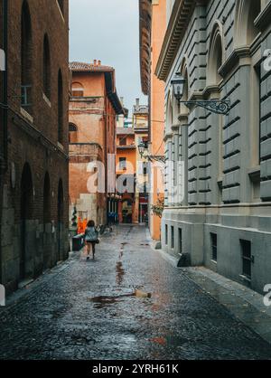 Die Frau geht vom Betrachter auf einer nassen Kopfsteinpflasterstraße weg, flankiert von historischen Gebäuden in der Stadt bologna, italien, nach dem Regen Stockfoto