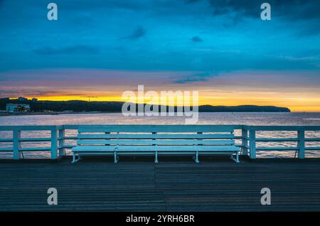 Die einladende weiße Bank steht am sopot Pier bei Sonnenaufgang und bietet einen friedlichen Blick auf den farbenfrohen Himmel und die ruhige ostsee Stockfoto