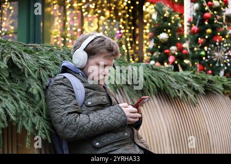 Mann in Kopfhörern sitzt mit Smartphone auf der Stadtstraße Neujahrsdekorationen Hintergrund, Winterferien Stockfoto