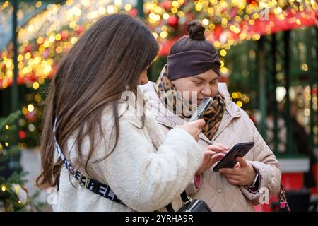 Zwei junge Frauen benutzen Smartphones auf der Stadtstraße auf Neujahrsdekorationen Hintergrund, Winterferien Stockfoto