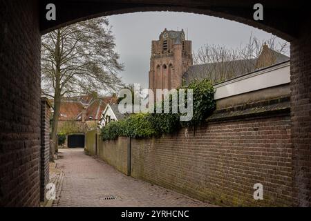 Eine kopfsteingepflasterte Gasse, die von Backsteinmauern eingerahmt ist, führt zu einem Blick auf einen historischen Kirchturm. Efeu-bedeckte Wände und malerische Häuser verleihen Charme unter einem bewölkten Himmel Stockfoto