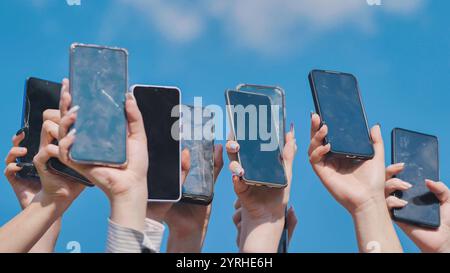 Viele Hände halten Smartphones mit gerissenen und kaputten Bildschirmen vor einem klaren blauen Himmel. Stockfoto