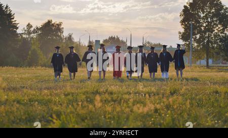 Eine Gruppe glücklicher Studenten, die Abschlusskleider tragen, läuft zusammen auf einem Feld bei Sonnenuntergang Stockfoto