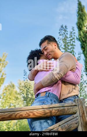 Ein liebevolles Paar umarmt sich herzlich auf einer Holzbrücke, umgeben von Bäumen und einem klaren blauen Himmel dieser intime Moment fängt Zuneigung, Verbindung und ein Stockfoto
