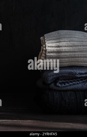 Ein kuscheliger Stapel gestrickter Kleidung in neutralen und dunklen Tönen. Die gefalteten Kleidungsstücke bieten vor dunklem Hintergrund Textur und Wärme, perfekt für fal Stockfoto
