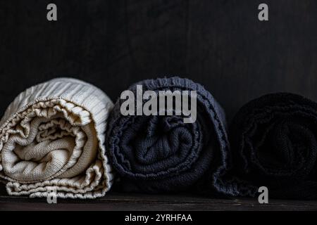 Drei kuschelige Strickpullover sind ordentlich gerollt und zeigen Texturen in Creme, Grau und Schwarz vor einem dunklen Hintergrund, die perfekt zur Illustration von Wärme geeignet sind Stockfoto