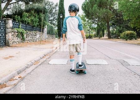 Ein Junge mit Helm Skateboards eine ruhige, von Bäumen gesäumte Straße hinunter Er trägt ein lässiges Outfit und kniehohe Socken und genießt ein Outdoor-Abenteuer auf einem friedlichen Weg Stockfoto