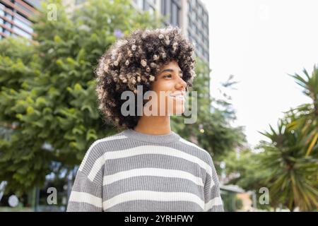 Eine fröhliche, gemischte Frau mit Vitiligo, lächelnd und genießt einen entspannenden Tag an der Universität, umgeben von üppigen grünen Bäumen auf einem Außengelände Stockfoto