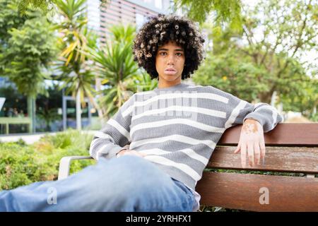 Gemischte Rassenfrau mit Vitiligo sitzt auf einer Bank auf einem Universitätscampus, umgeben von Grün, sie genießt einen friedlichen und entspannenden Moment im Freien Stockfoto