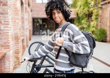Eine gemischte Frau mit Vitiligo steht mit ihrem Fahrrad und lächelt zufrieden auf einem ruhigen Universitätscampus. Sie hält einen Rucksack und strahlt ein Gefühl von Relevanz aus Stockfoto
