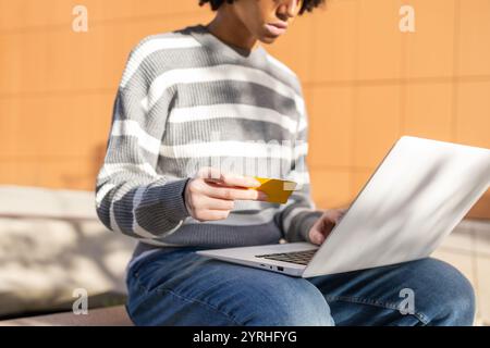 Eine gemischte Frau mit Vitiligo sitzt auf dem Campus mit einem Laptop und einer Kreditkarte. Sie ist lässig in einem gestreiften Pullover und Jeans gekleidet Stockfoto