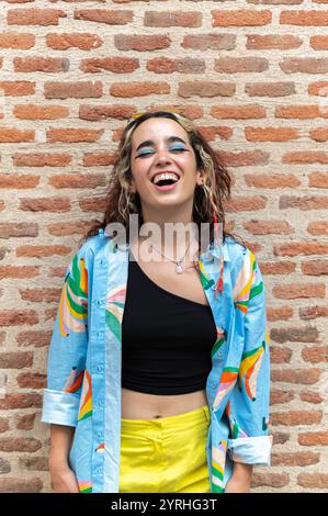 Ein lebhaftes Porträt einer fröhlichen jungen Frau mit lockigem Haar, die von Herzen lacht. Sie trägt ein farbenfrohes Hemd und eine gelbe Hose, steht gegen ein R Stockfoto