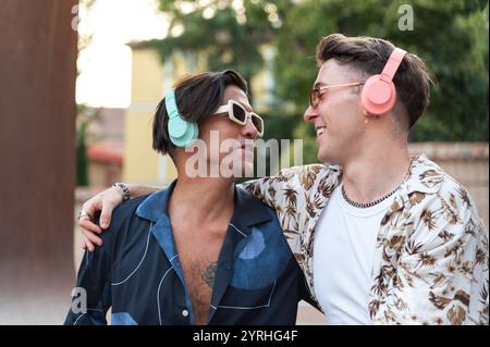Eine fröhliche Szene von zwei jungen Männern in stilvollen Outfits, die bunte Kopfhörer tragen und draußen einen warmen, freundlichen Look teilen. Stockfoto
