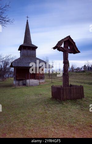 Rustikale Holzkirche und Kreuz in ländlichen Maramures, eingebettet in Rumäniens Karpaten, die Szene fängt traditionelle Architektur und heiteres Cu ein Stockfoto