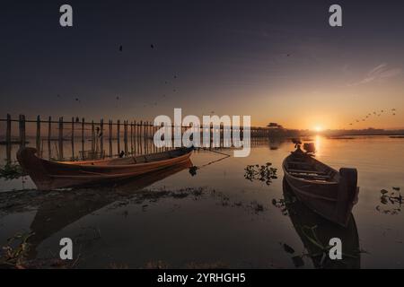 Ein ruhiger Sonnenaufgang über einem ruhigen See mit einem hölzernen Pier im Hintergrund und zwei traditionellen Booten im Vordergrund, umgeben von sanften Wellen und bir Stockfoto