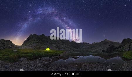 Ein glühendes Zelt unter der Milchstraße, eingerahmt von zerklüfteten Bergen und einem Sternenhimmel. Die Reflexion in einem ruhigen Teich trägt zur ruhigen, atemberaubenden Nacht bei Stockfoto