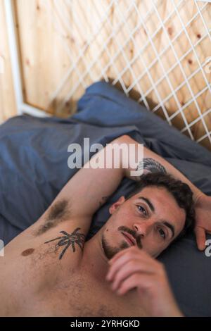 Ein Mann mit komplizierten Tätowierungen liegt bequem auf einem Bett mit blauen Laken, umgeben von Holzwänden und einem weißen Gitter. Die Szene vermittelt Entspannung Stockfoto