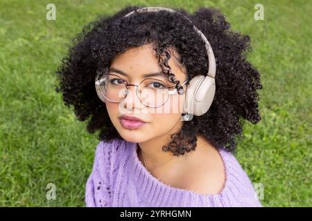 Junge Afro-Latino-Frau mit Kopfhörern und einem lila Pullover, die auf grünem Gras sitzt, ihre lockigen Haare und Brillen spiegeln einen entspannten, modernen Lebensstil wider Stockfoto