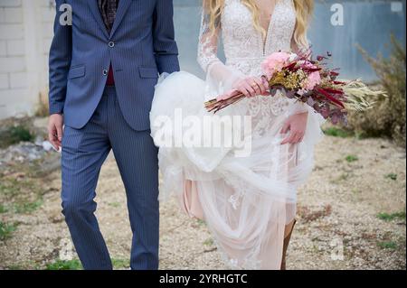 Eine Nahaufnahme einer Braut in einem Spitzenkleid mit Blumenstrauß, begleitet von einem Bräutigam in einem modernen Marineblau-Anzug, fängt die Essenz der stilvollen Hochzeit fa ein Stockfoto