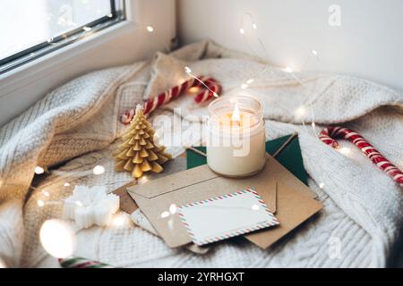 Warme Weihnachtsszene am Fenster mit einer leuchtenden Kerze, festlichen Umschlägen, Zuckerstangen und einem kleinen dekorativen Baum auf einer Strickdecke, die an eine gemütliche Atmosphäre erinnert Stockfoto