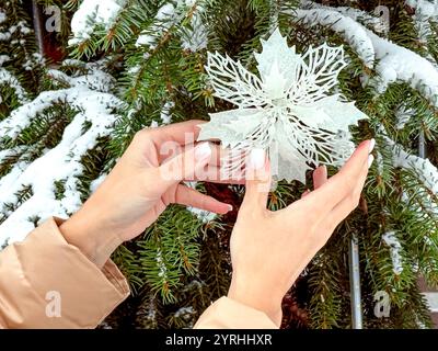 Mit den Händen legen Sie eine funkelnde Schneeflockendekoration vorsichtig auf eine schneebedeckte Kiefer und fangen den festlichen Geist der Weihnachts- und Neujahrsvorbereitungen ein Stockfoto