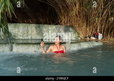 Eine asiatische Frau in rotem Bikini macht ein Selfie, während sie sich in den natürlichen Thermalwässern der Terme di Saturnia, Toskana, Italien, inmitten üppiger Fol, entspannt Stockfoto