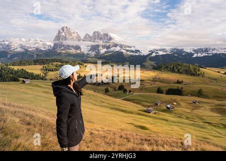 Eine Frau in einer schwarzen Jacke zeigt in Richtung der majestätischen Dolomiten, umgeben von den weiten Wiesen der Seiser Alm, Italien schneebedeckten Gipfeln und scatt Stockfoto