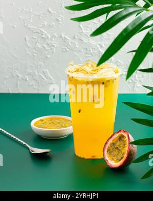 Eine lebendige Tasse eisgekühlten Passionsfruchtsaft, begleitet von geschnittenem Obst und einer kleinen Schüssel Fruchtfleisch, die an einer strukturierten Wand und grünem Laub für ein T T Stockfoto