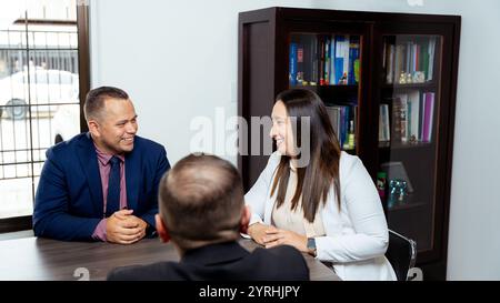 Drei formell gekleidete Latino-Geschäftsleute, die an einem Meeting teilnehmen, sitzen an einem Tisch mit einem Schrank voller Bücher im Hintergrund Stockfoto