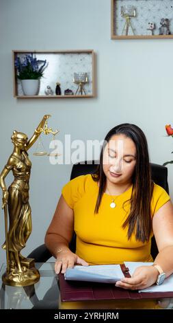 Eine Anwältin in gelbem Outfit prüft Dokumente an ihrem Schreibtisch, die mit einer Lady Justice Statue geschmückt sind, die Gerechtigkeit symbolisiert, und gesetzliche Regale mit Dekor sind im Th Stockfoto