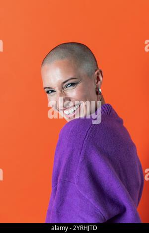 Eine fröhliche, kahlköpfige Erwachsene Frau in einem purpurroten Pullover posiert vor einem leuchtenden orangefarbenen Hintergrund. Ihr warmes Lächeln strahlt Zuversicht und Glück aus Stockfoto