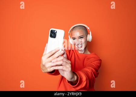 Eine fröhliche, kahlköpfige Erwachsene Frau in einem orangefarbenen Sweatshirt macht ein Selfie, während sie weiße Kopfhörer trägt und sich vor einem orangefarbenen Hintergrund posiert. Stockfoto