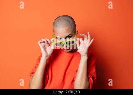 Eine kahlköpfige Erwachsene Frau trägt ein leuchtendes orangefarbenes Oberteil und passt eine übergroße grüne Sonnenbrille auf einen passenden orangefarbenen Hintergrund an Stockfoto