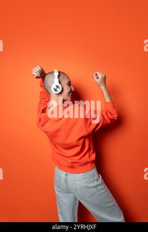 Eine kahlköpfige Erwachsene Frau in einem leuchtenden orangefarbenen Hoodie und einer grauen Jeans tanzt fröhlich vor einem orangefarbenen Hintergrund und genießt Musik über weiße Kopfhörer. Stockfoto