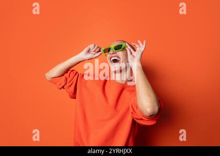 Fröhliche Erwachsene Frau in einem orangefarbenen Hemd, die ihre leuchtend grüne Neon-Sonnenbrille auf einem passenden orangefarbenen Hintergrund verspielt anpasst. Ihre Begeisterung Stockfoto