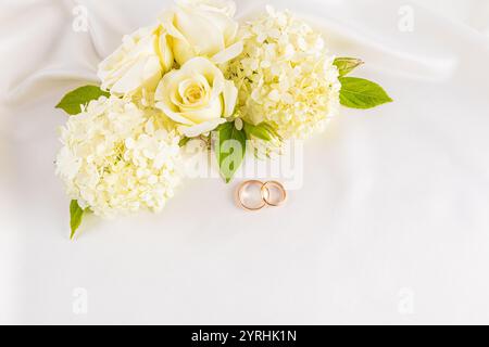 Zwei Eheringe ruhen elegant auf einem weichen Satinhintergrund, begleitet von einem zarten Bouquet aus weißen Rosen und Hortensien, perfekt für eine rom Stockfoto