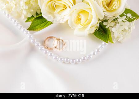 Zwei elegante Eheringe ruhen auf einem luxuriösen Satin-Stoff, begleitet von zarten weißen Blumen und einer Perlenschnur, ideal für Romantik und Hochzeit Stockfoto